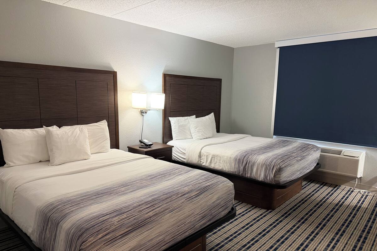 A hotel room with two beds, a nightstand with a lamp, a window with a view, and a radiator.