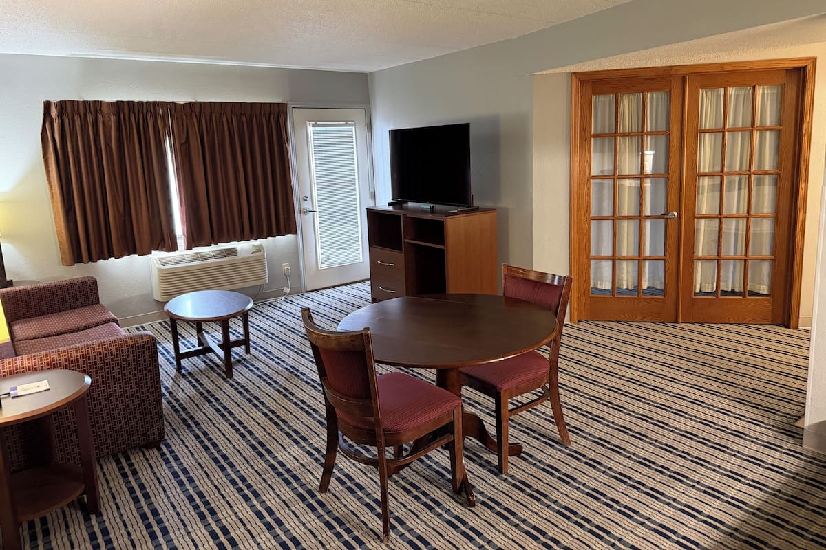 A hotel room with a dining table, chairs, a sofa, a TV, and a large window with curtains.