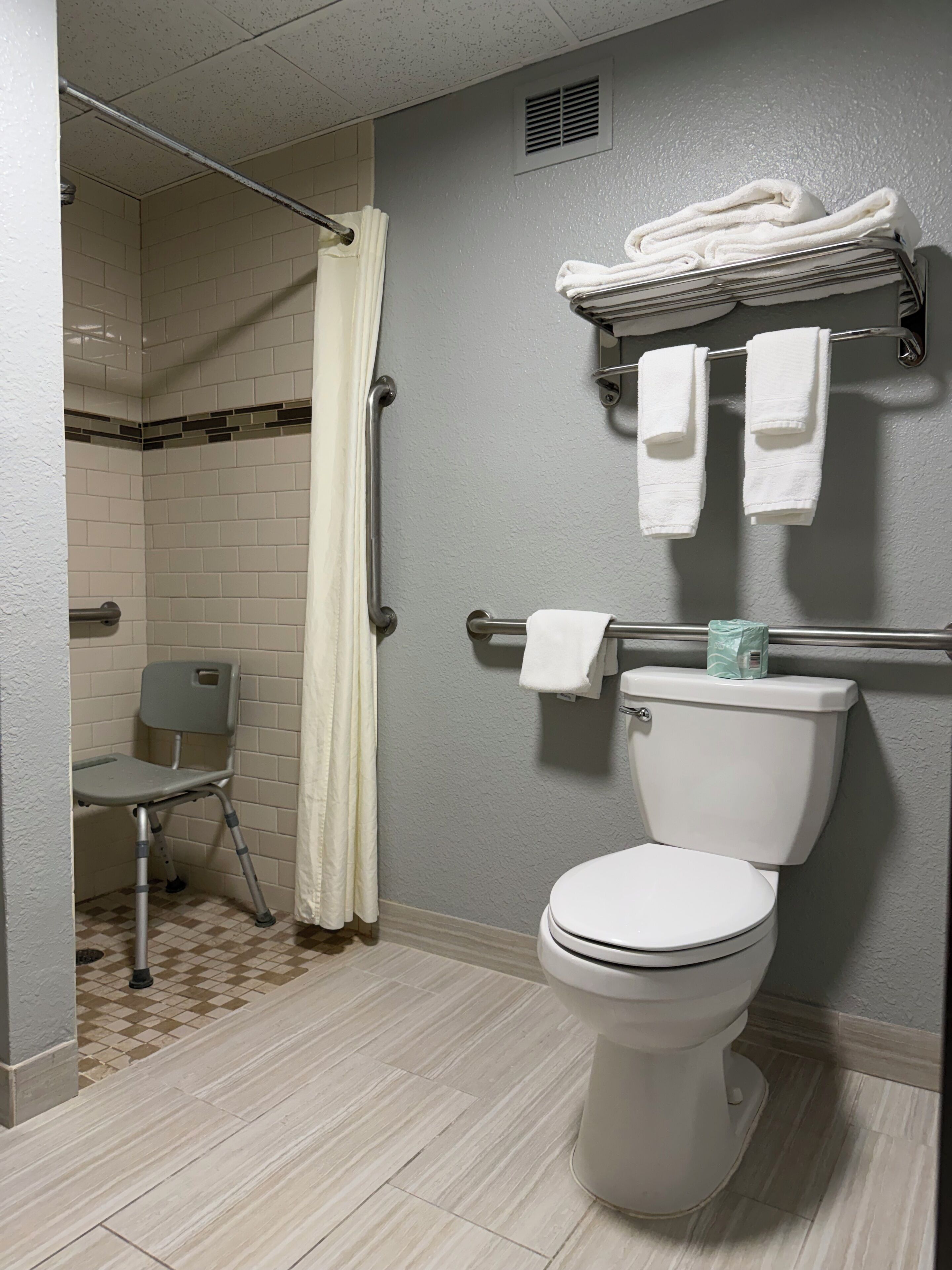 A bathroom with a toilet, a shower with a curtain, a grab bar, a chair, and a towel rack with towels.