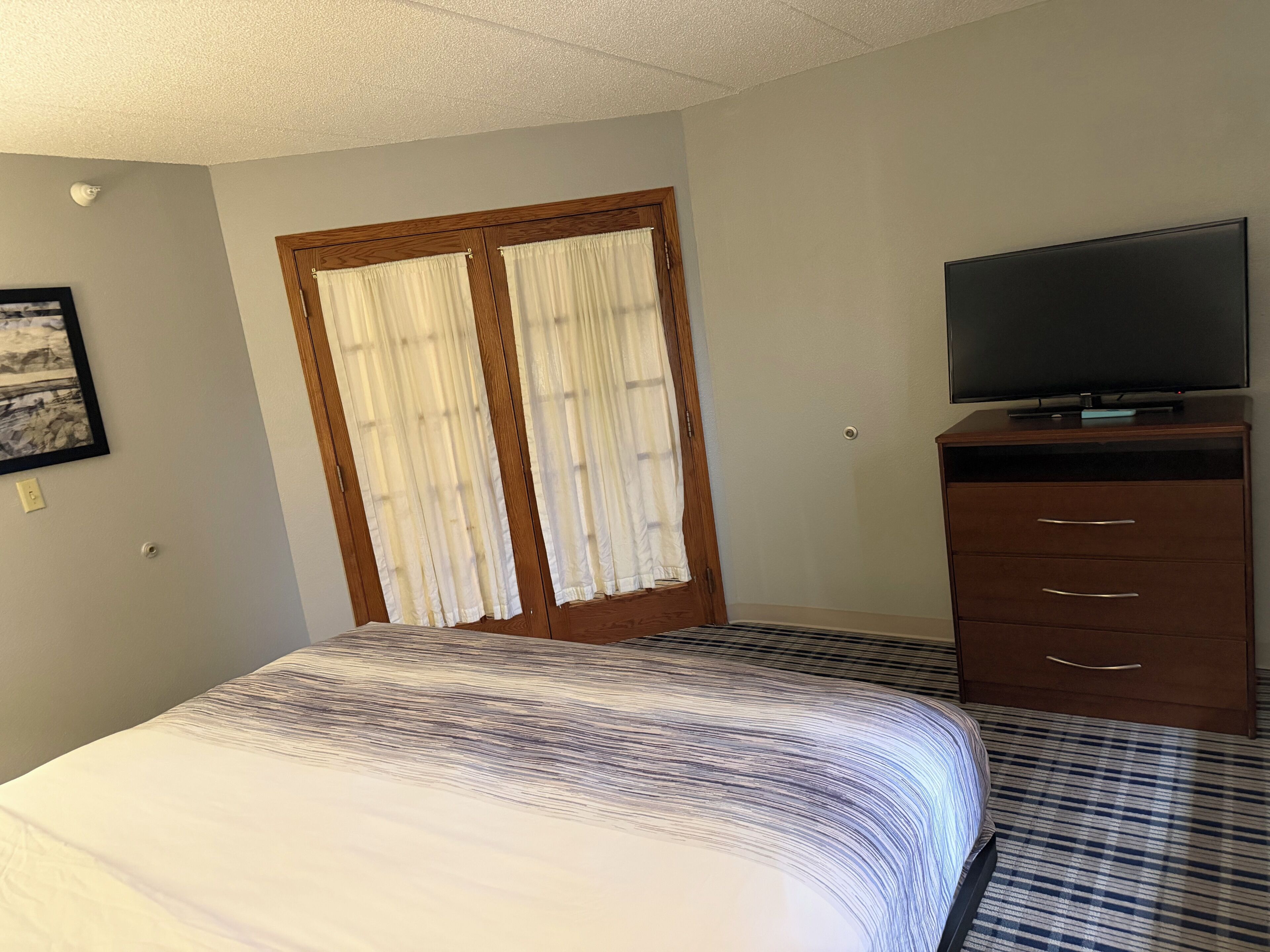 A hotel room with a bed, a television on a wooden dresser, and a door with curtains.