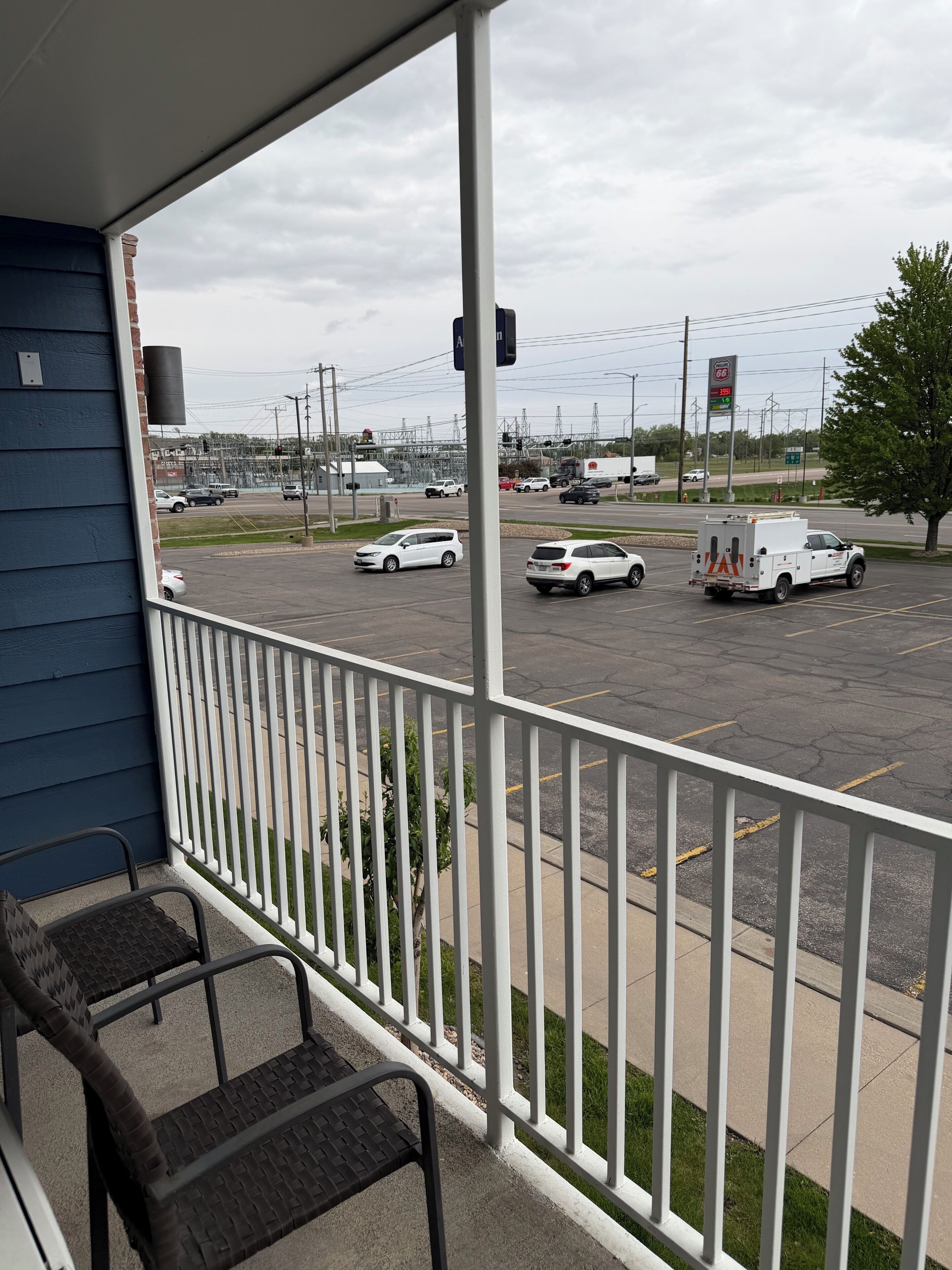 A balcony with a railing, a view of a parking lot with cars and a utility truck, and a building with a blue exterior in the background.