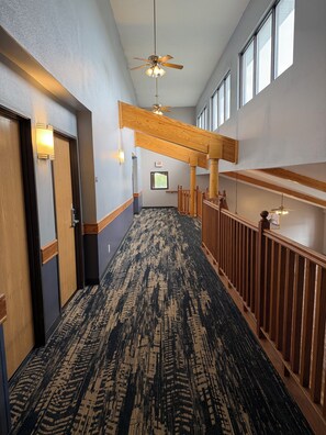 A hotel hallway with a patterned carpet, wooden railings, and ceiling fans. - AmericInn by Wyndham Kearney (Kearney)