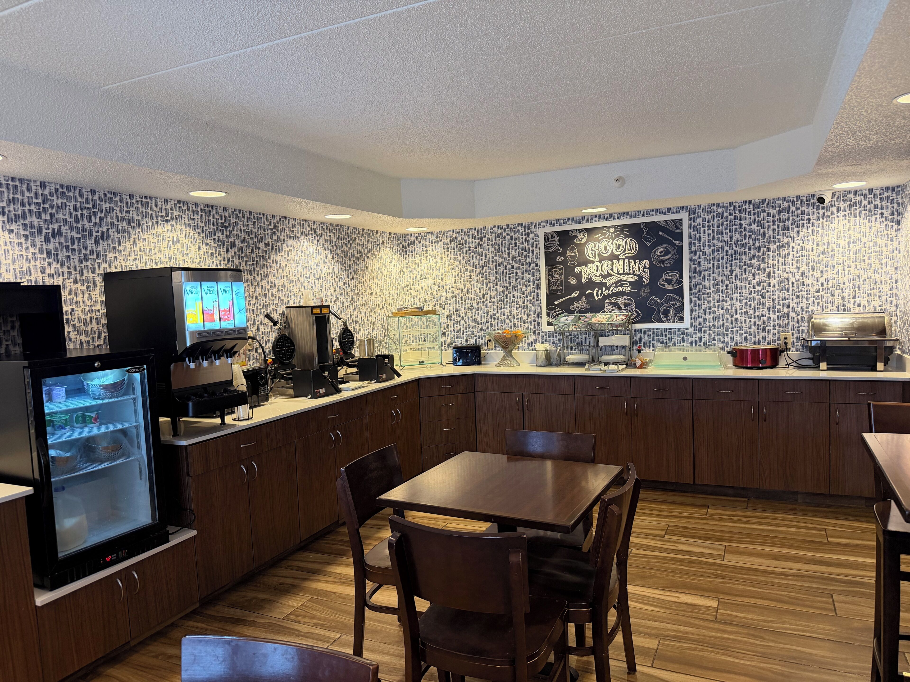 A kitchen area with a coffee machine, a refrigerator, and a chalkboard with a 'Good Morning' message.