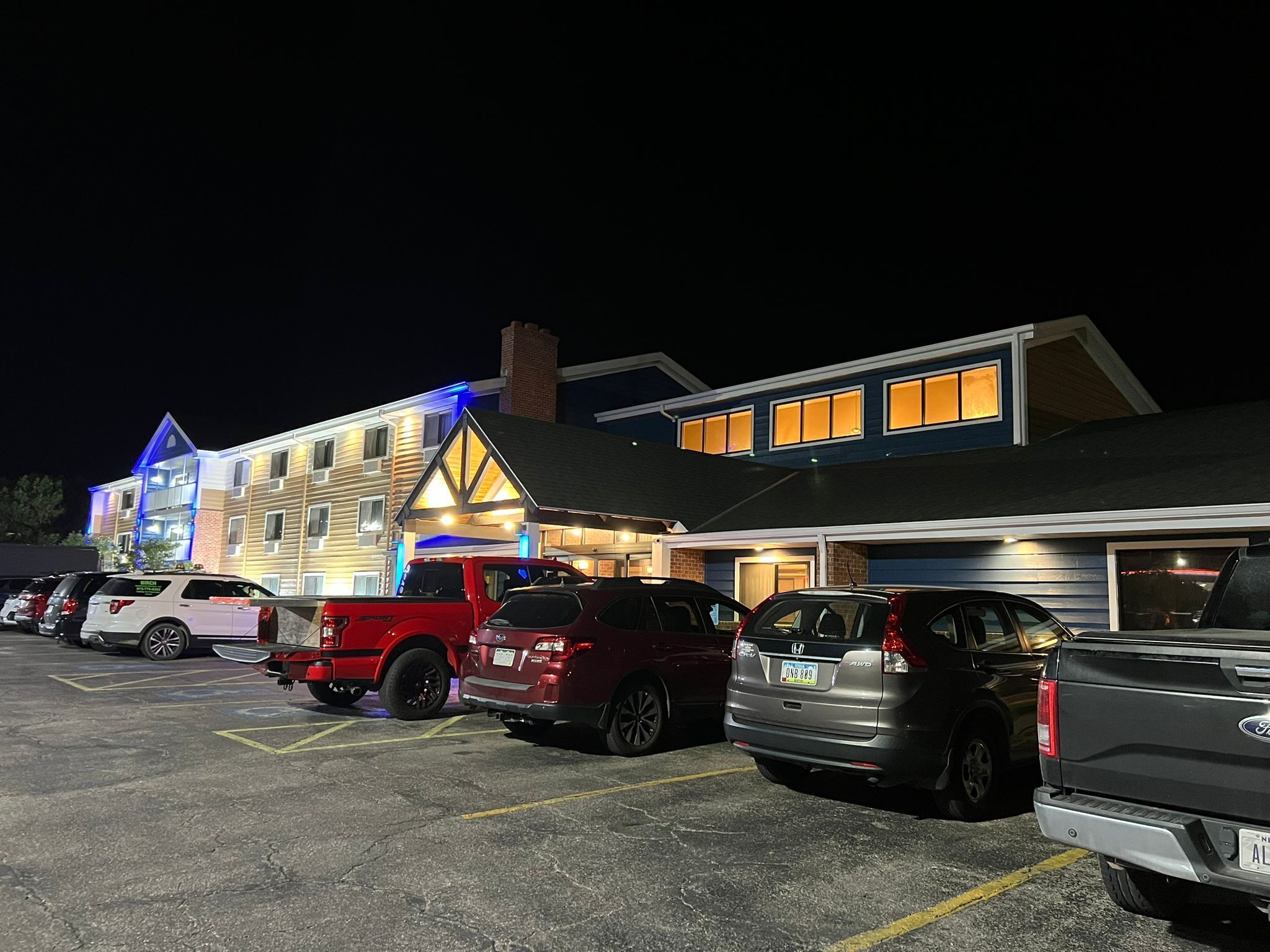 Um estacionamento com vários carros e um edifício com janelas iluminadas e entrada com iluminação azul.