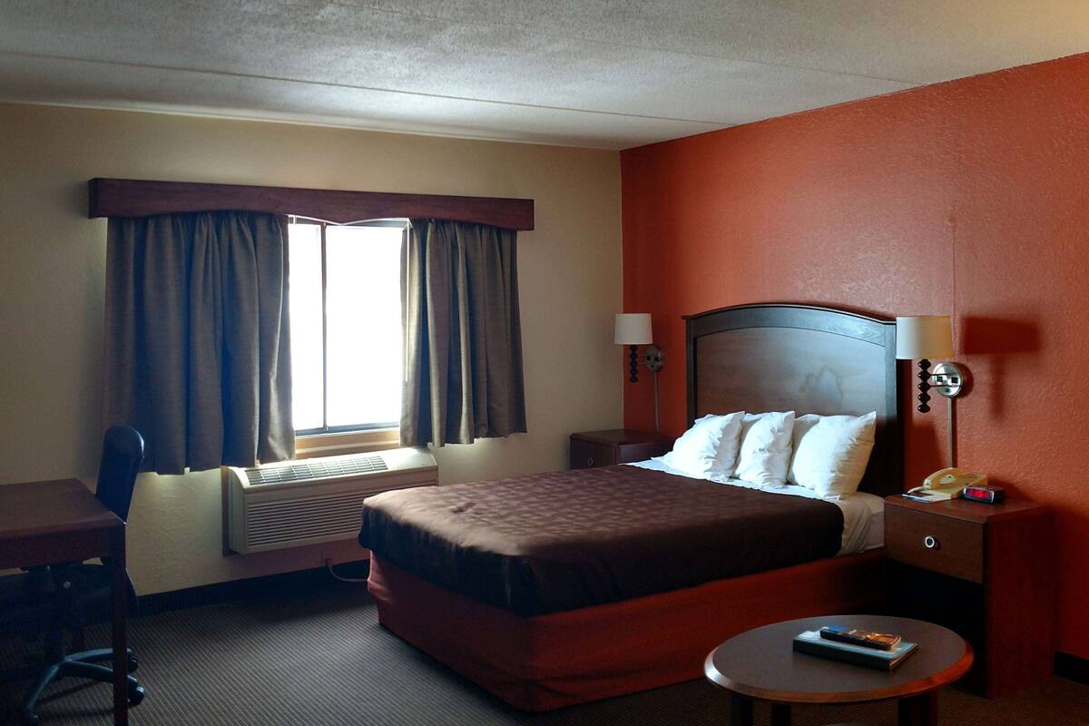 A hotel room with a bed, a desk, a chair, a bedside table with a lamp, and a window with curtains.