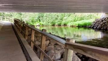 Une allée couverte, dotée d’une rambarde en bois, surplombe une rivière paisible, le tout étant entouré de verdure.
