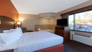Habitación de hotel con cama, escritorio, televisor de pantalla plana, ventana con vista a coches aparcados y árboles, y baño con bañera y toallero.