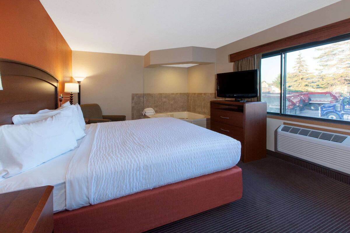 A hotel room with a bed, a desk, a flat-screen TV, a window with a view of parked cars and trees, and a bathroom with a bathtub and a towel rack.