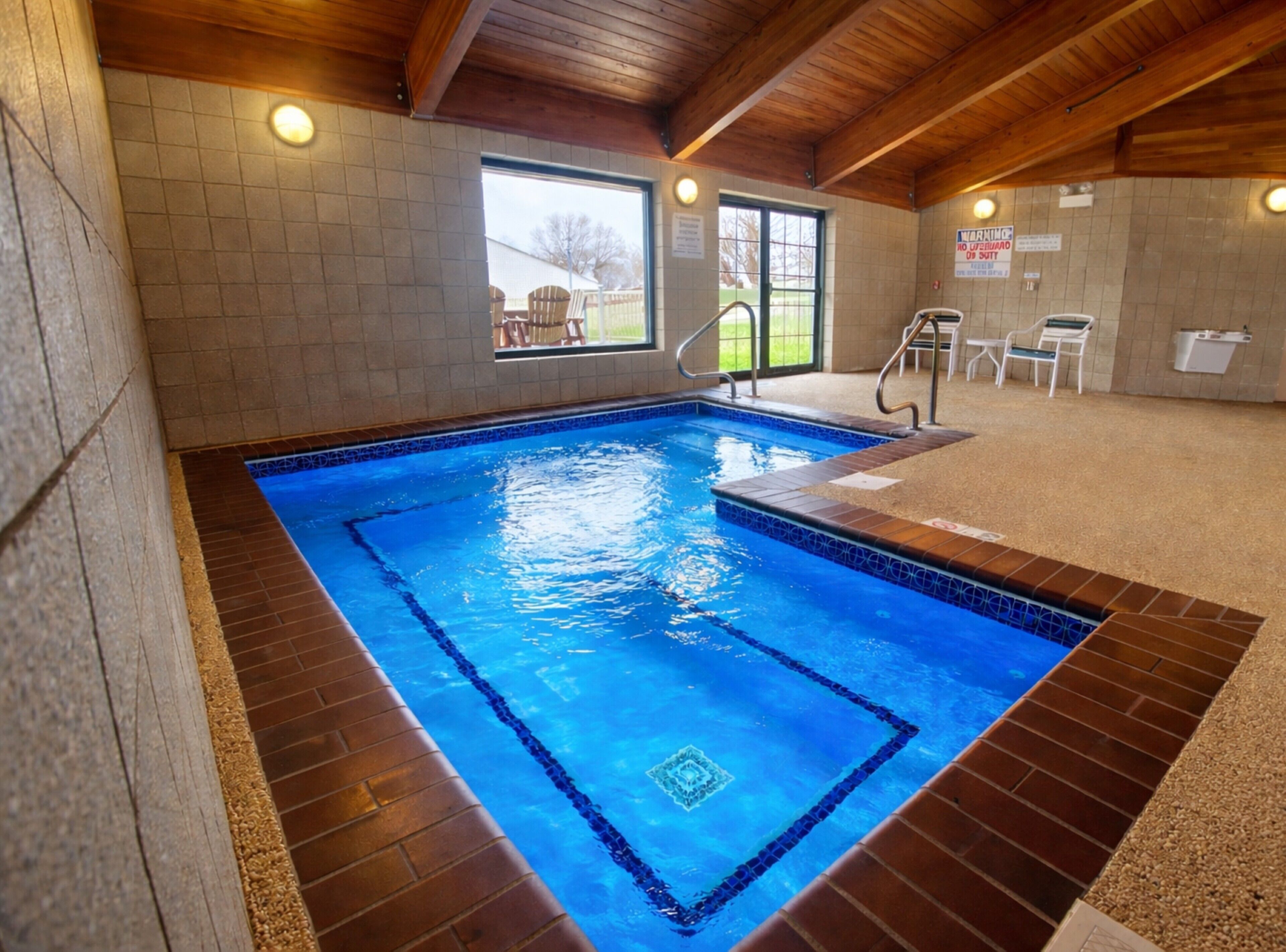 En indendørs swimmingpool med fliserede vægge, trælofter og udsigt til græsset udenfor.