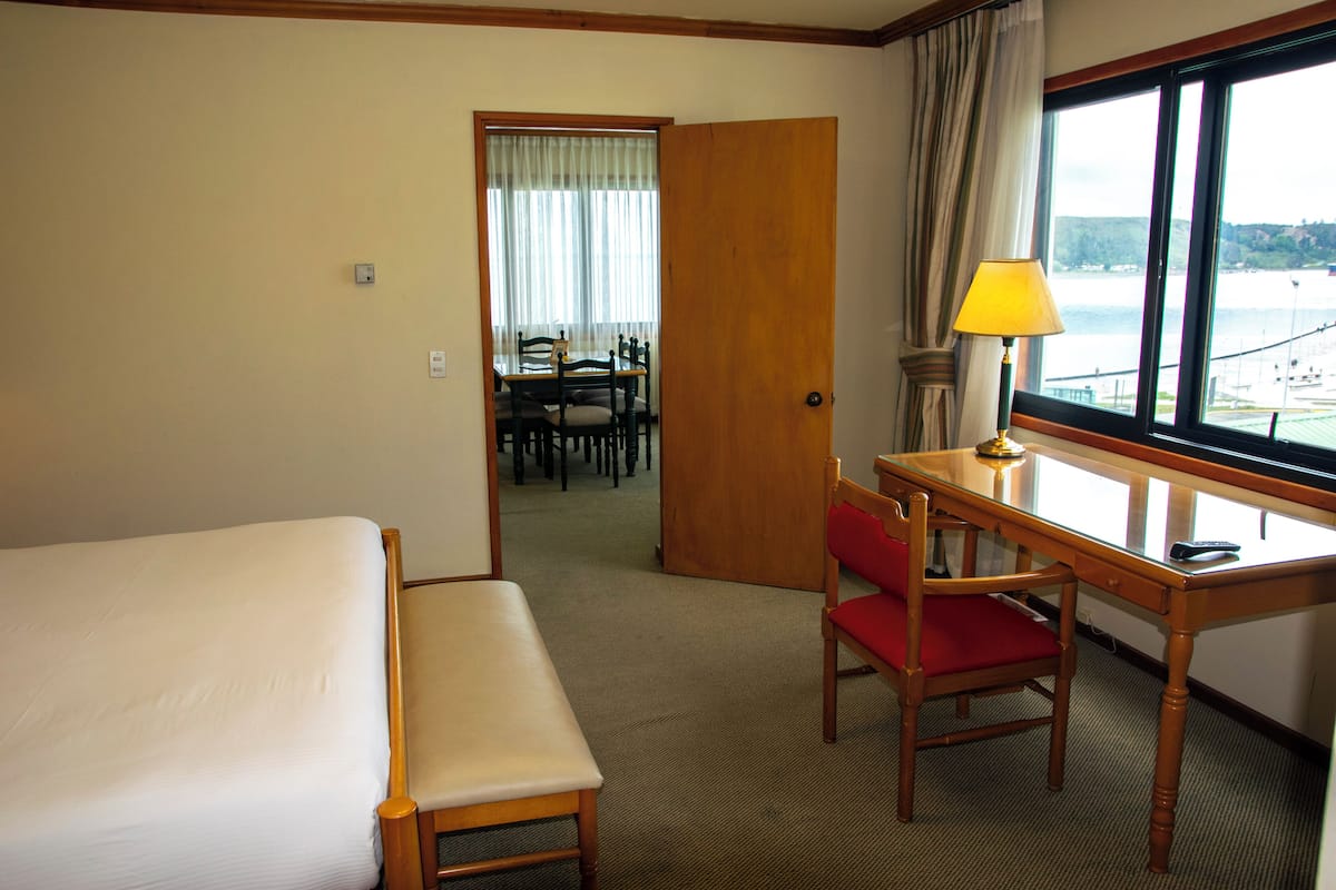 A hotel room with a bed, a desk with a chair, a lamp, and a view of a body of water through a window.