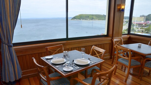 Comedor con mesa dispuesta para cuatro, sillas de madera con cojines azules y vistas al mar a través de amplios ventanales.