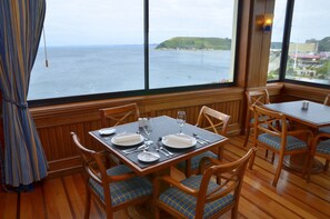 A dining area with a table set for four, wooden chairs with blue cushions, and a view of the sea through large windows. - Abba Presidente Suites Puerto Montt (Puerto Montt)