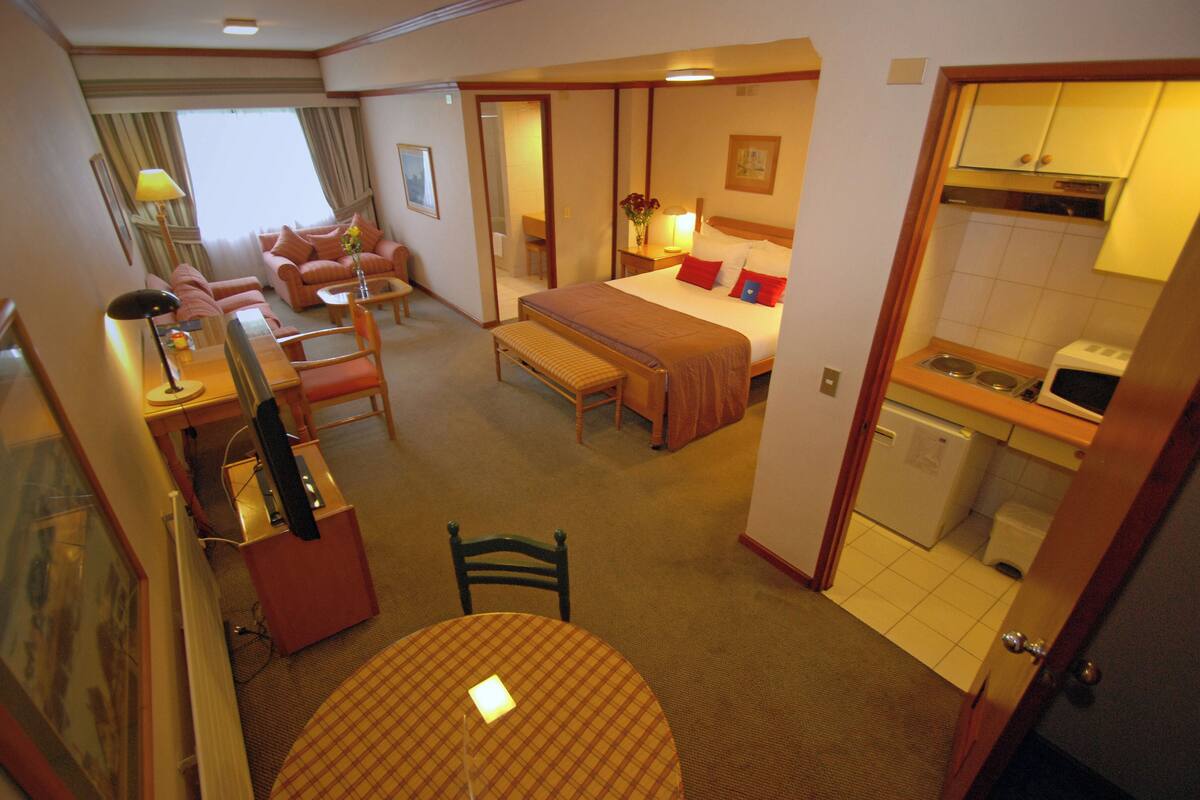 A hotel room with a bed, a desk, a chair, a table, a sofa, a small kitchen area, and a bathroom.