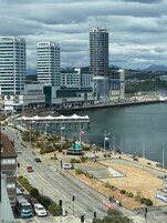 Paisaje urbano frente al agua con edificios modernos, un puerto deportivo y una zona peatonal con una gran escultura.