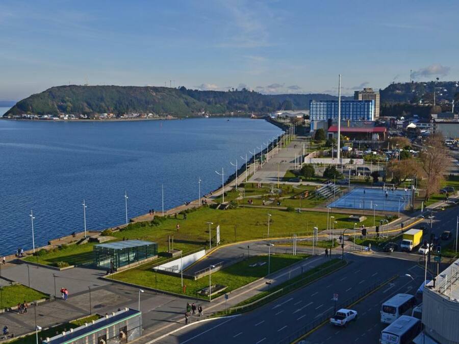Eine Küstenstadt mit einem markanten Gebäudekomplex, einer Uferpromenade und einer Straße, die zu einer Brücke führt.
