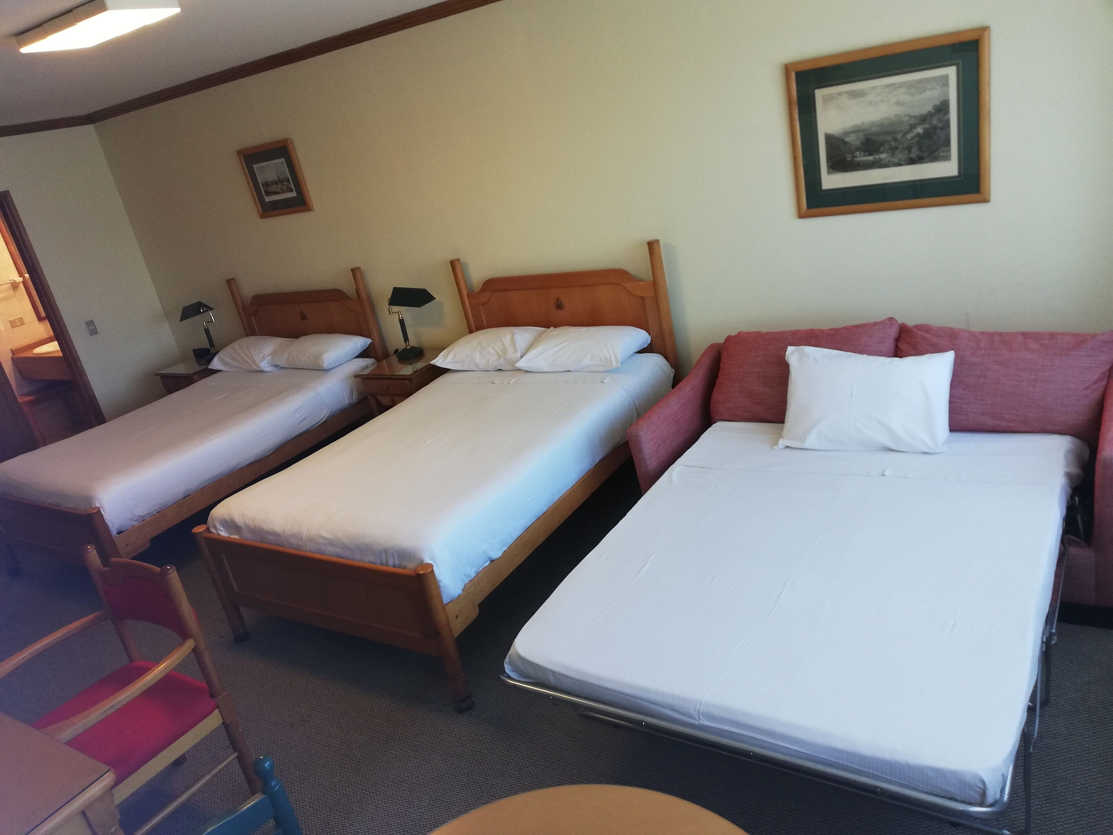 Une chambre d’hôtel avec trois lits simples, une tête de lit en bois, un canapé rouge et un tableau encadré au mur.
