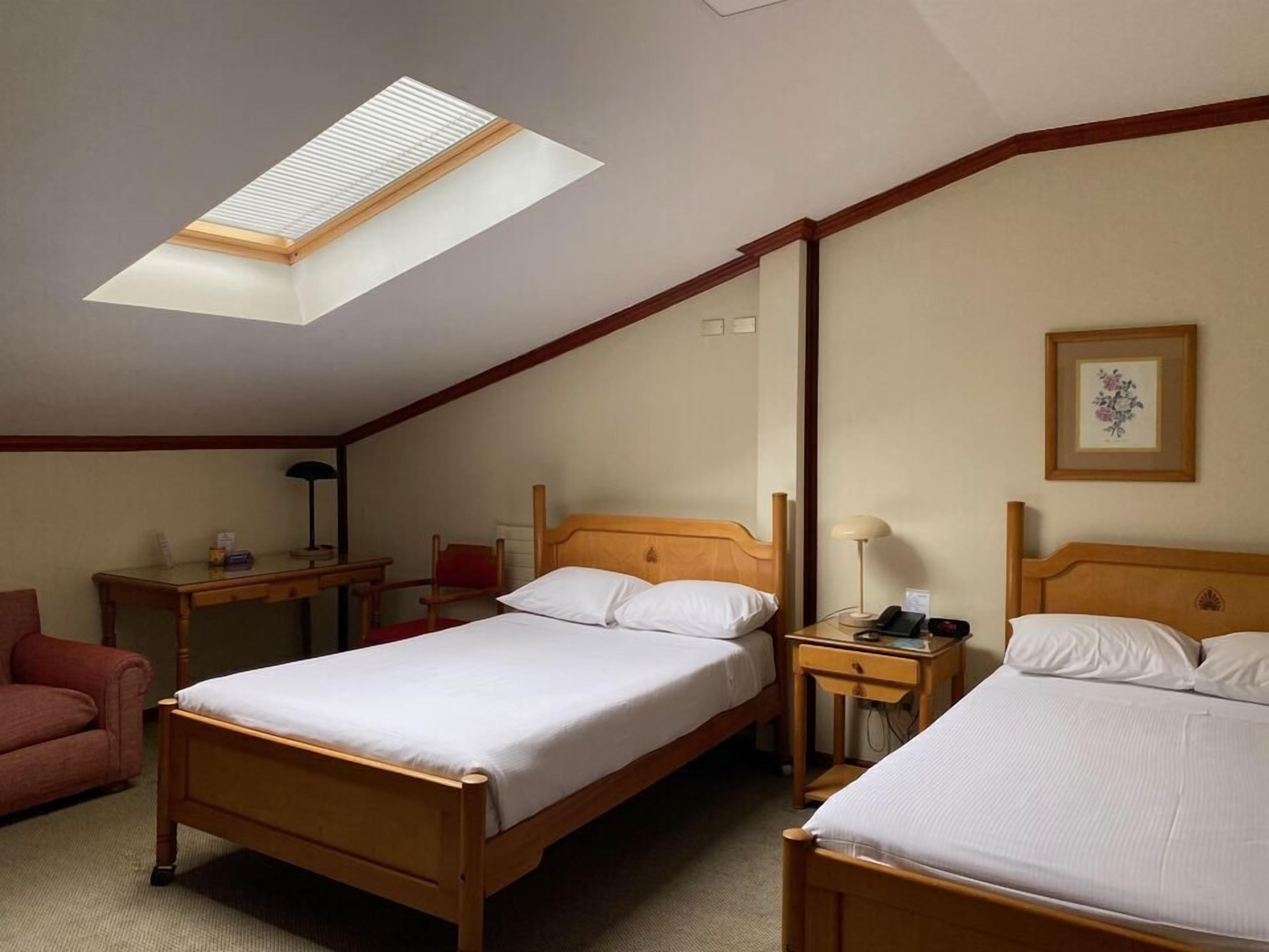 Habitación de hotel con dos camas de madera, un escritorio, una silla, una mesita y un tragaluz.
