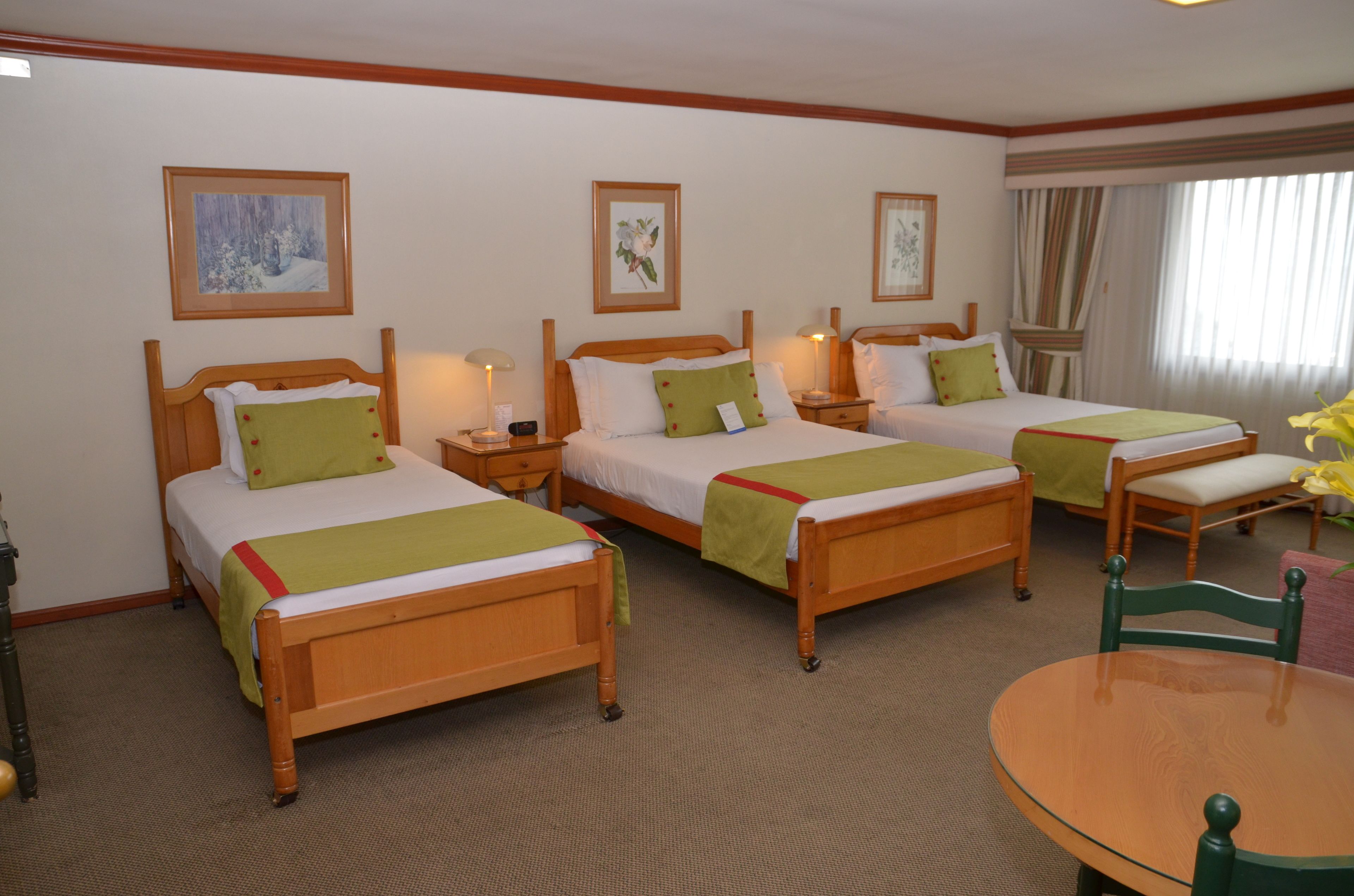A hotel room with three wooden beds, each with a green blanket and white bedding. There are bedside tables with lamps, a round wooden table, and a chair. The room has a carpeted floor and a window with curtains.