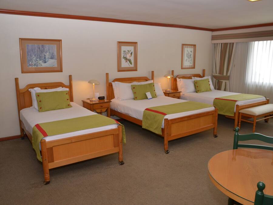 A hotel room with three wooden beds, each with a green blanket and white bedding. There are bedside tables with lamps, a round wooden table, and a chair. The room has a carpeted floor and a window with curtains.