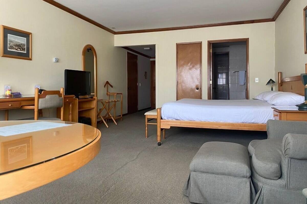 A hotel room with a bed, a desk with a computer, a television, a chair, a small table, and a door leading to another room.