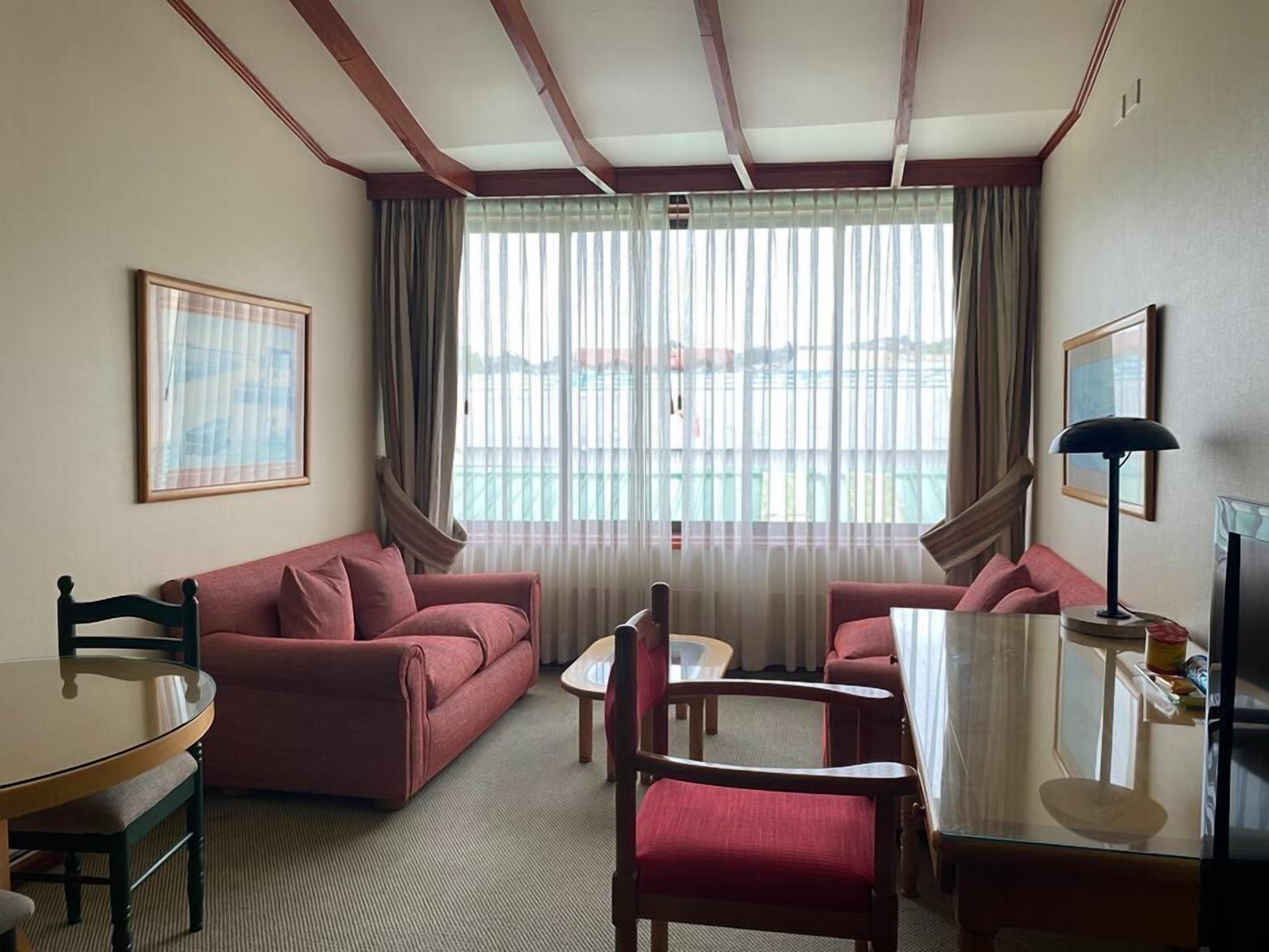 A hotel room with a red sofa, a wooden chair, a small table, and a lamp. There is a window with curtains and a view of the outside.