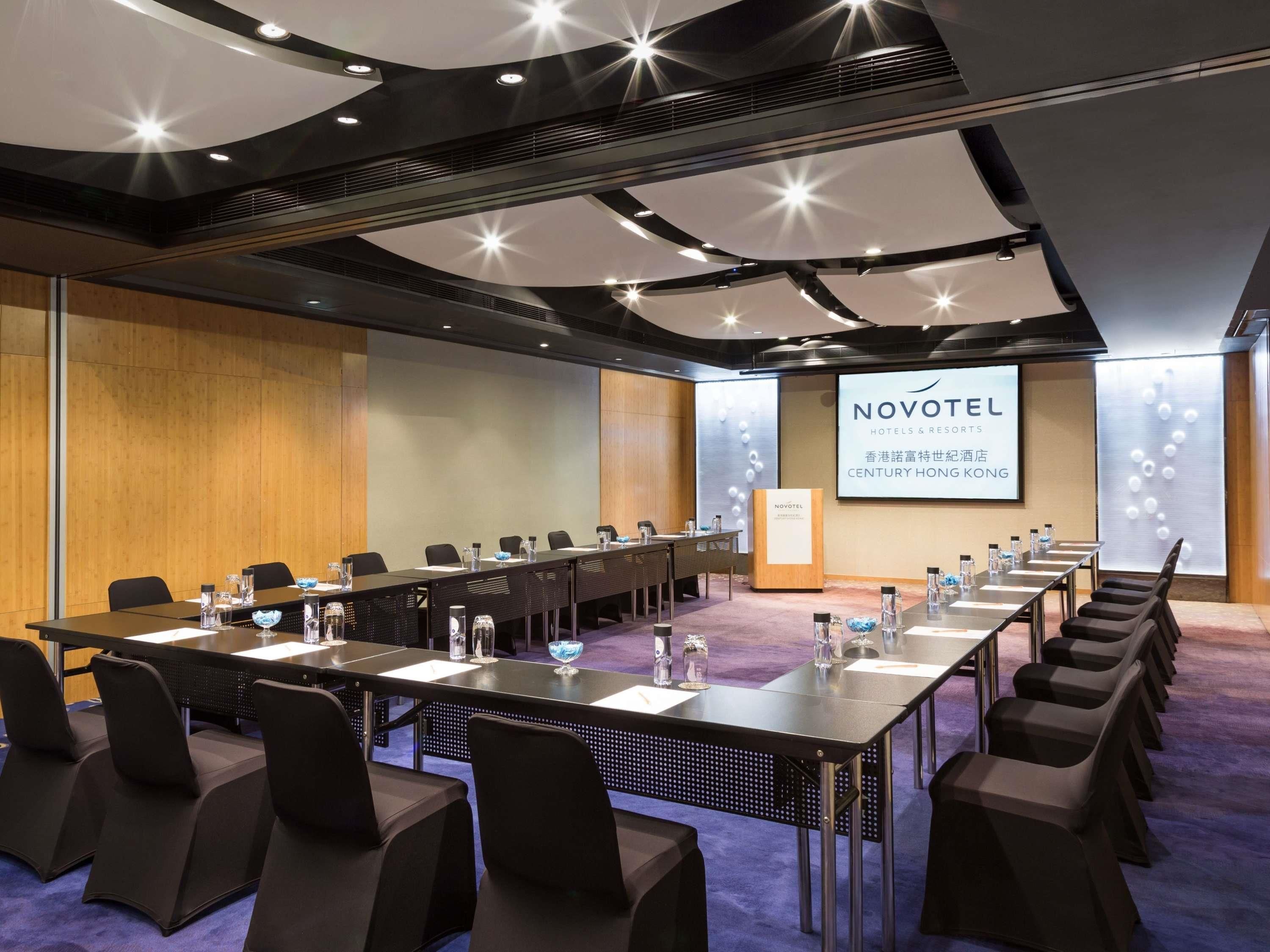 A conference room with a long table, chairs, a podium, and a screen displaying 'NOVOTEL HOTELS & RESORTS CENTURY HONG KONG'.