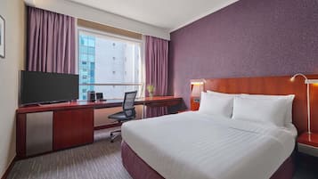 A hotel room with a large bed, a desk with a flat-screen TV, a chair, and a desk lamp.