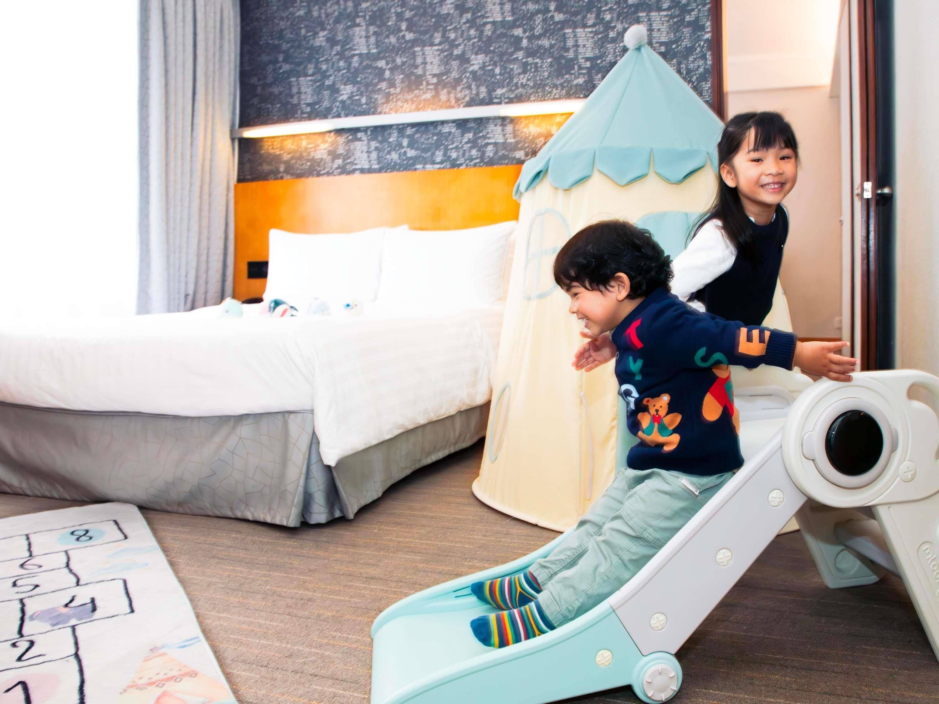 A child on a slide in a hotel room with a bed, a play mat, and a wall with a textured design.