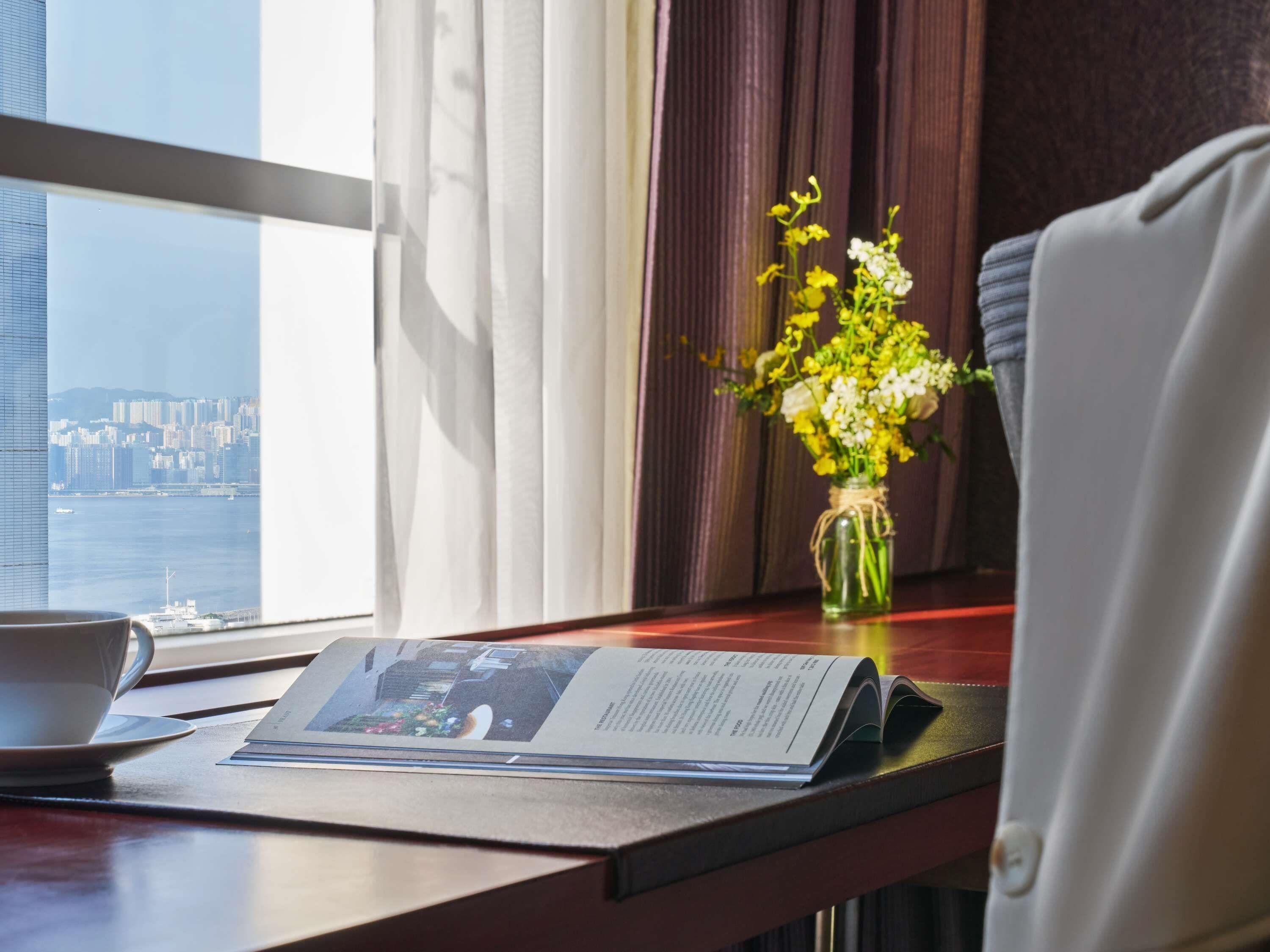Habitación de hotel con vista a un paisaje urbano, una mesa con periódico, un florero con flores y una silla.