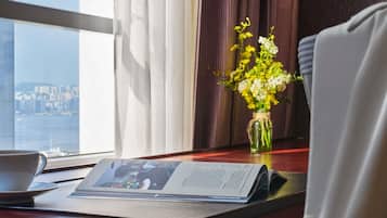 Habitación de hotel con vista a un paisaje urbano, una mesa con periódico, un florero con flores y una silla.