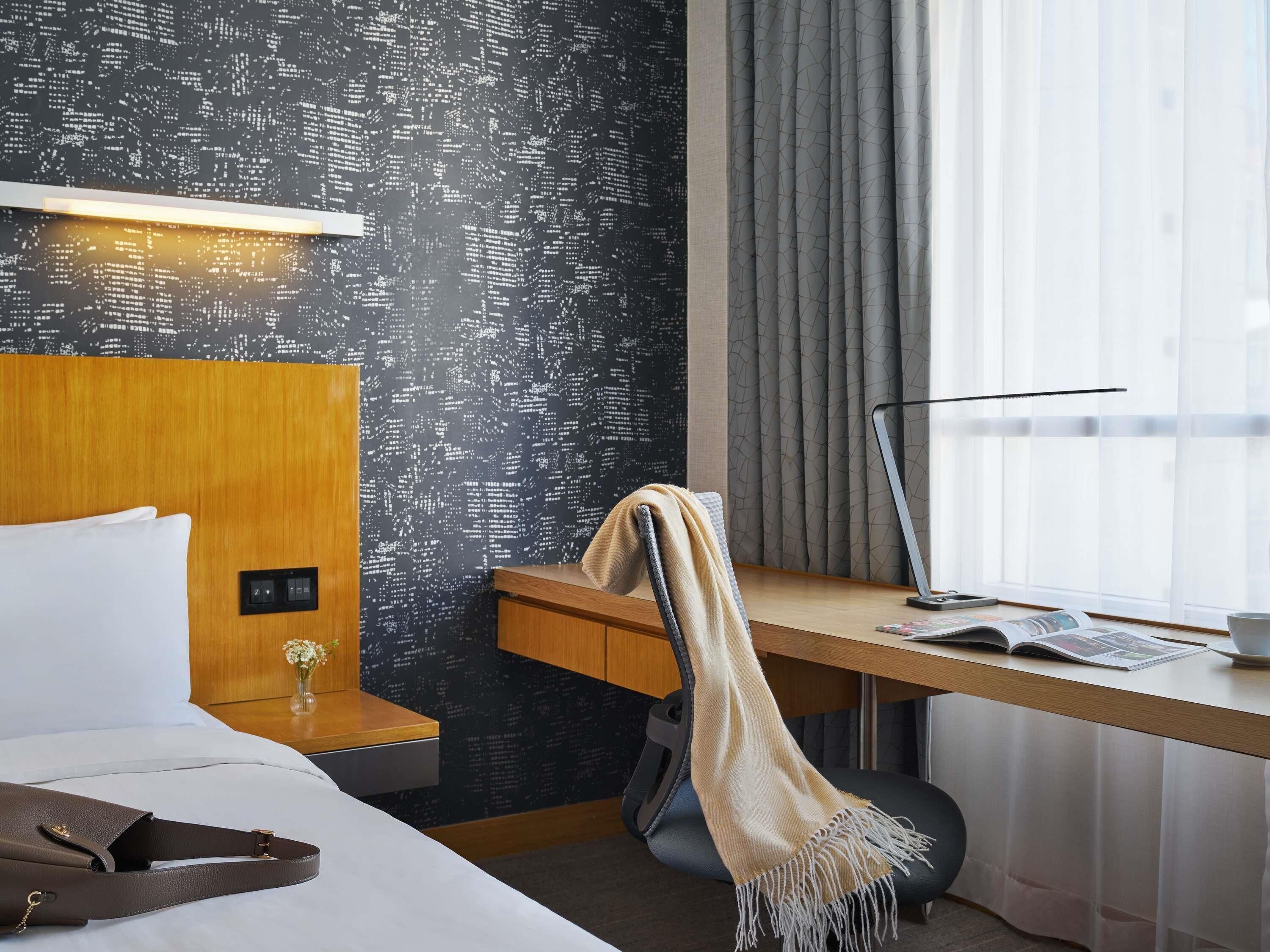 Hotelkamer met een bed, een bureau met lamp, een stoel, een raam met gordijnen en een wand met een stadsgezicht patroon.