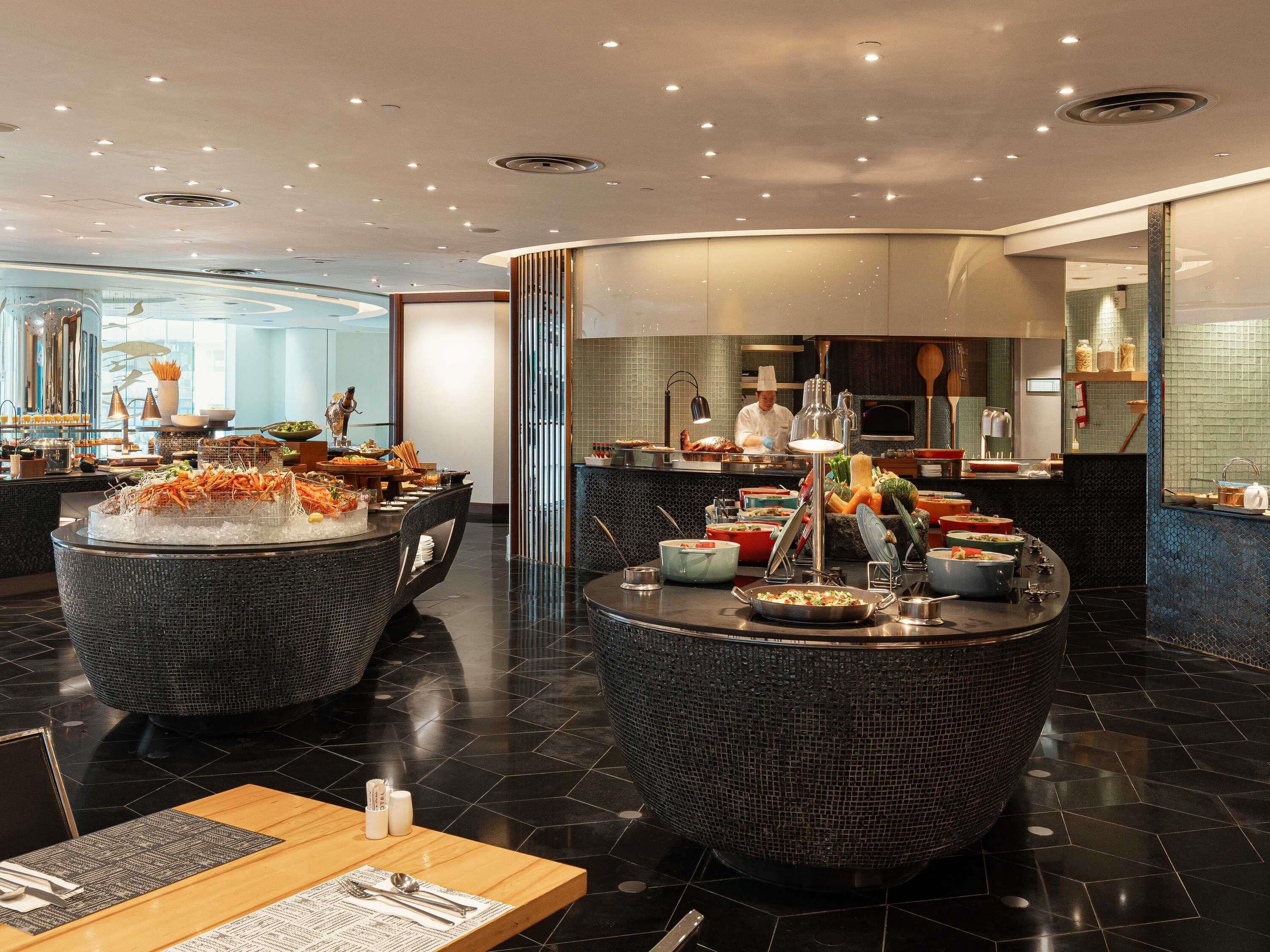 A modern hotel lobby with a central dining area featuring round tables with food displays and a kitchen in the background.