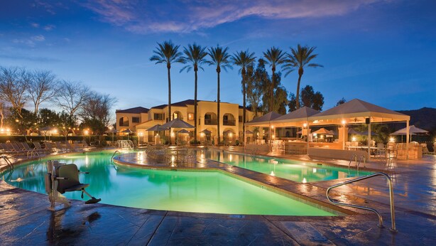 A swimming pool with lounge chairs and umbrellas, surrounded by palm trees and a building with multiple balconies.