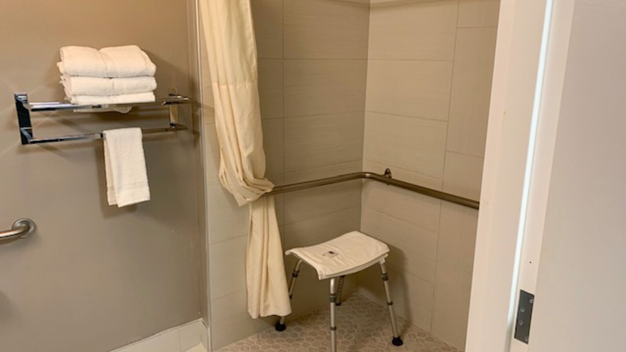 A small, enclosed shower area with a glass door, a shower curtain, a towel rack with folded towels, and a small step stool.