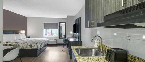 A compact hotel room with a kitchenette, a bed with patterned bedding, a small desk, and a window providing natural light.