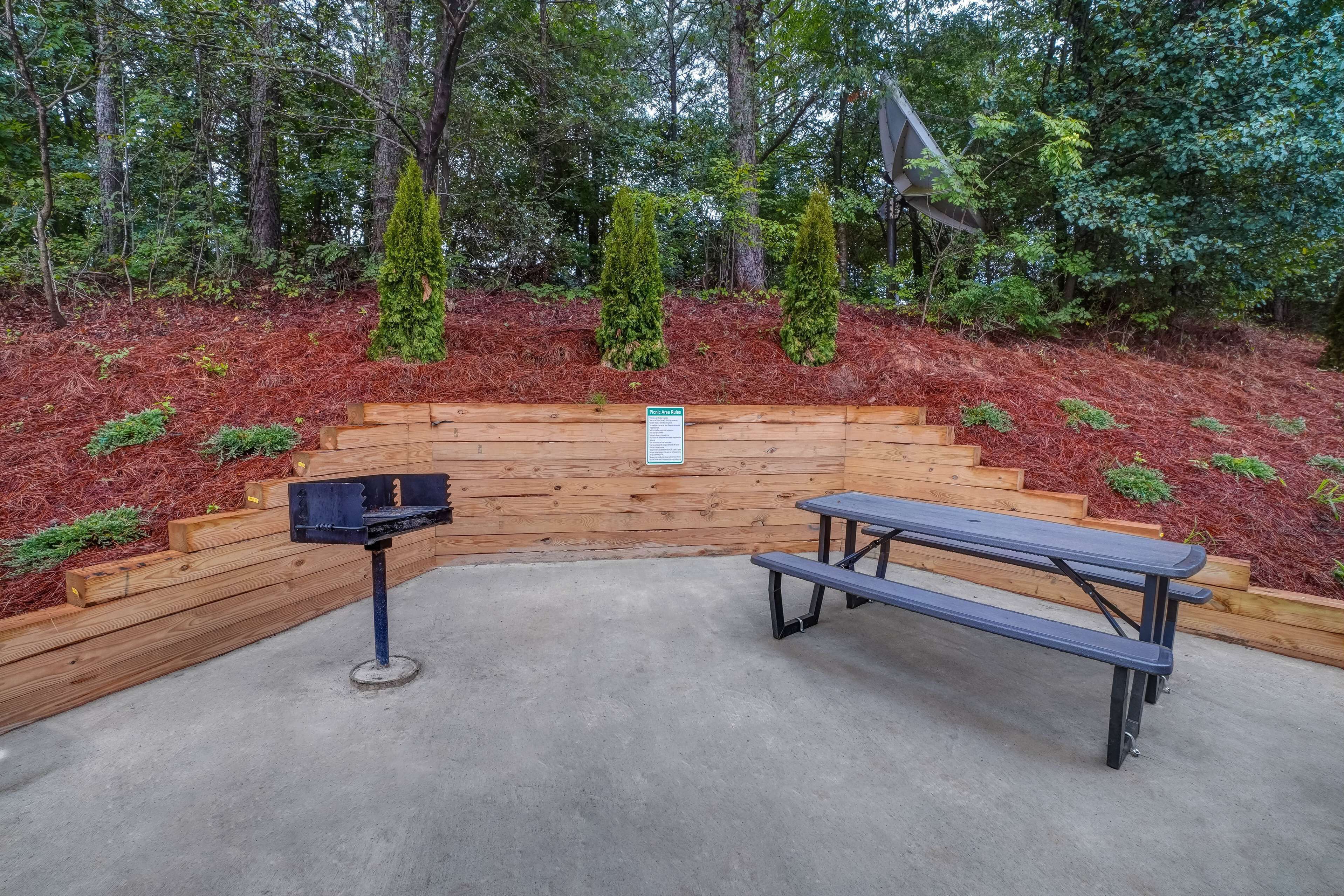 A picnic area with a bench and grill, surrounded by a wooden fence and mulch, with trees in the background.