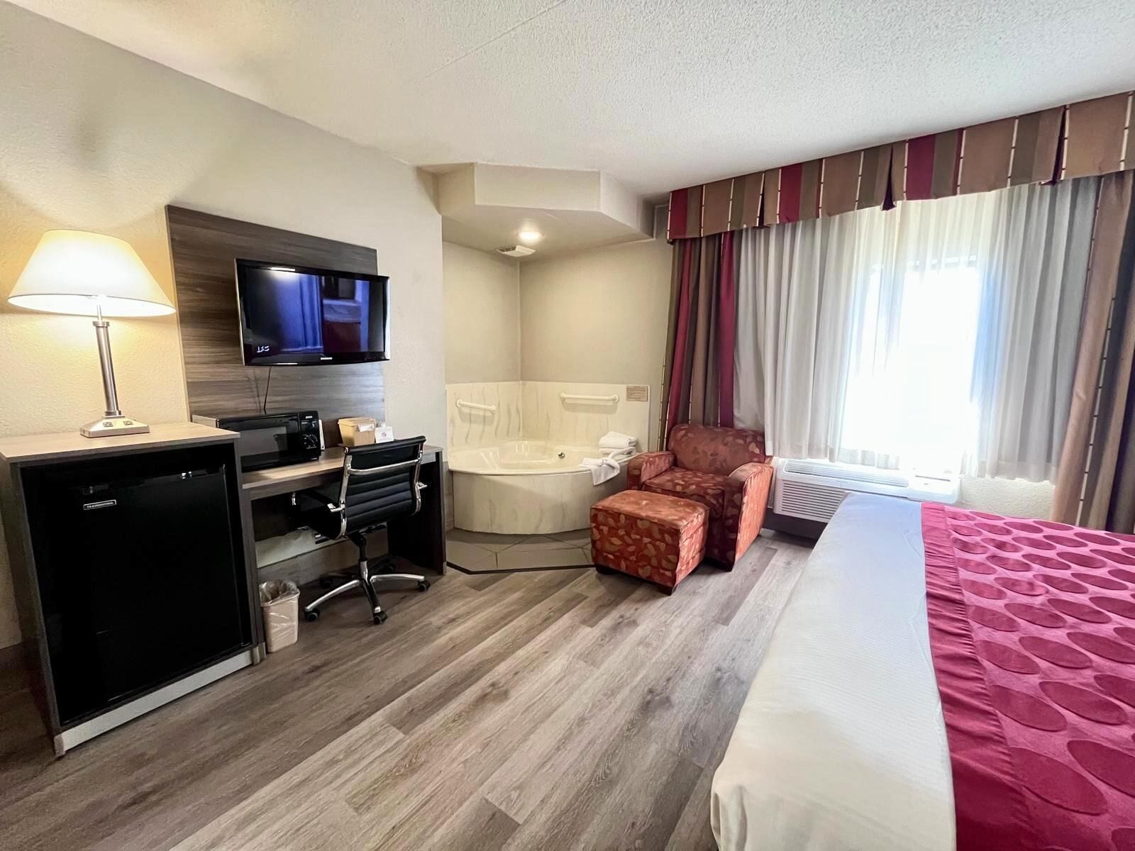 A hotel room with a bed, a desk with a lamp, a TV, a small table, a chair, a sofa, a bathtub, and a window with curtains.