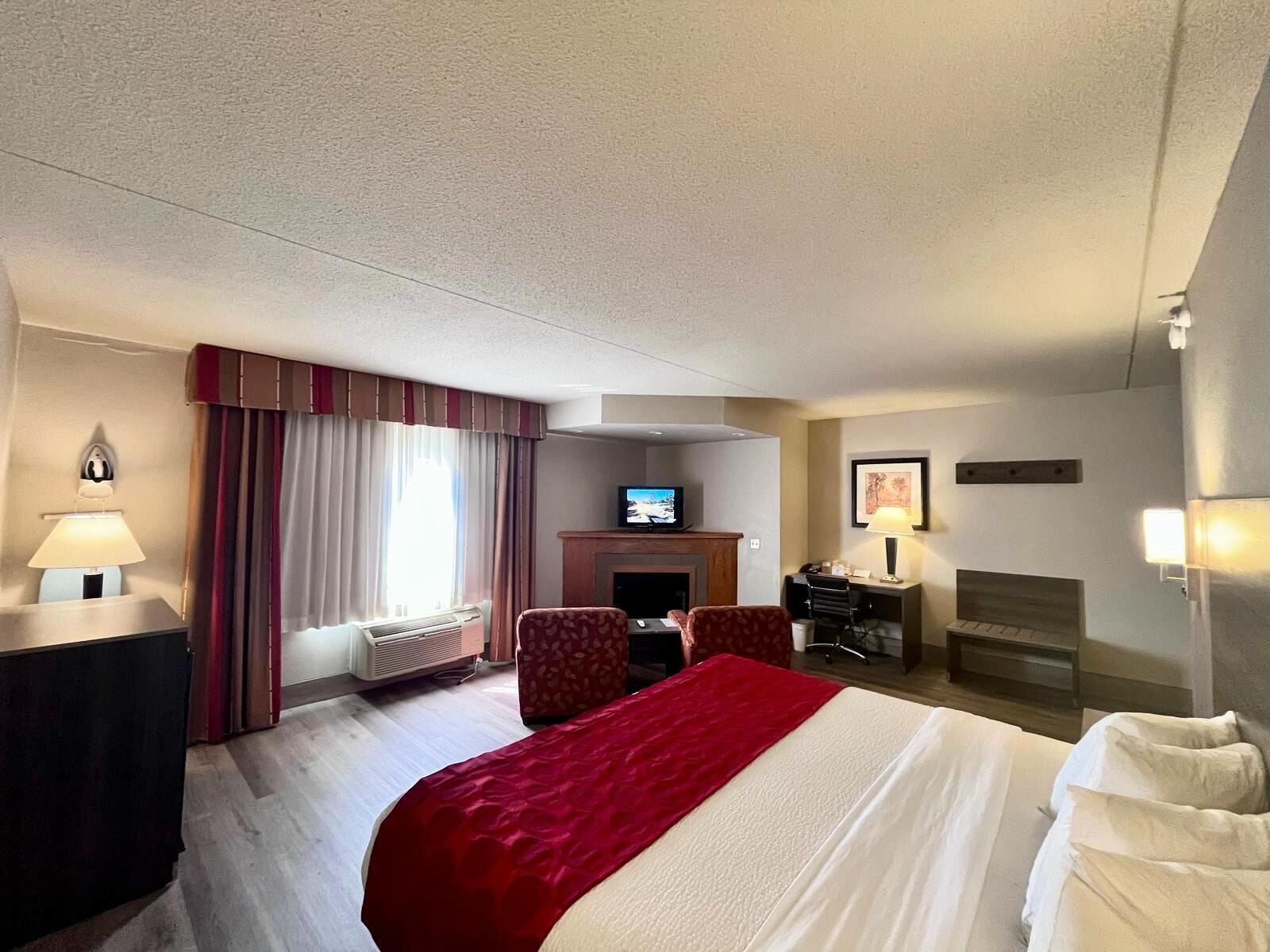 A hotel room with a large bed, a desk, a chair, a television, and a fireplace.
