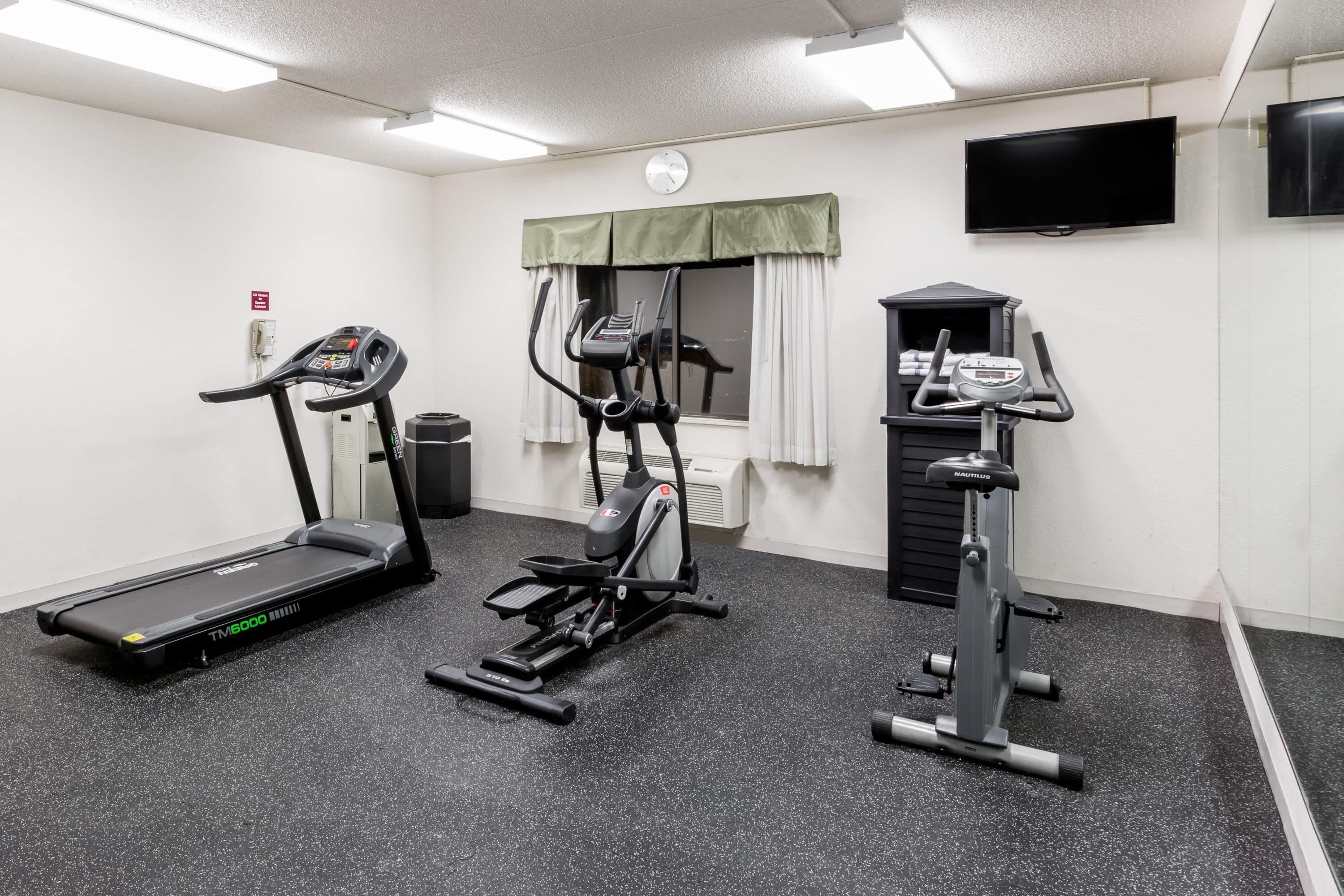 Un gimnasio con caminadora, elíptica y bicicleta estática, un reloj y un televisor en la pared.