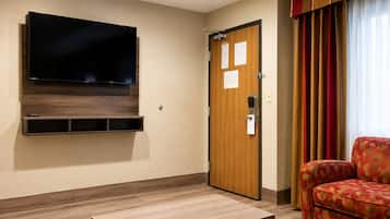 A hotel room with a flat-screen TV mounted on the wall, a wooden door with a lock, a red patterned sofa, and a wooden coffee table.