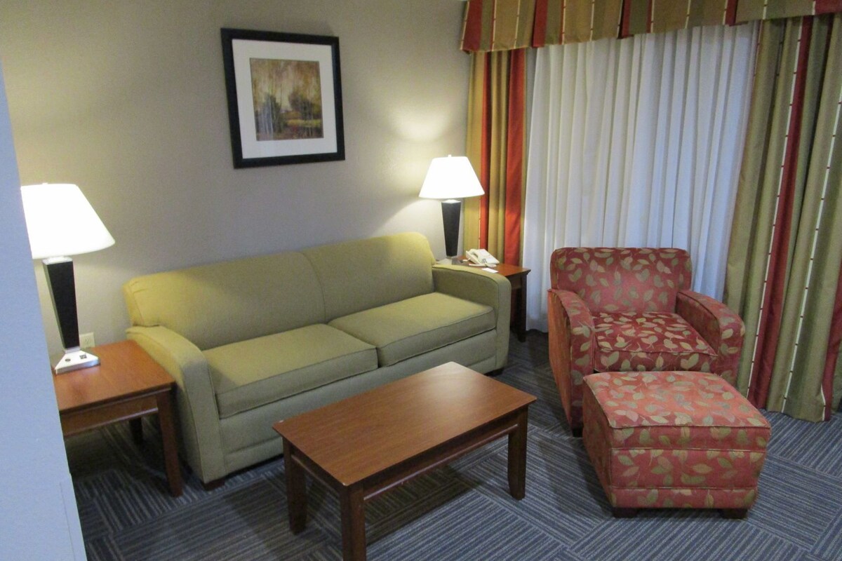 A hotel room with a sofa, armchair, ottoman, table, lamps, and a framed picture on the wall.