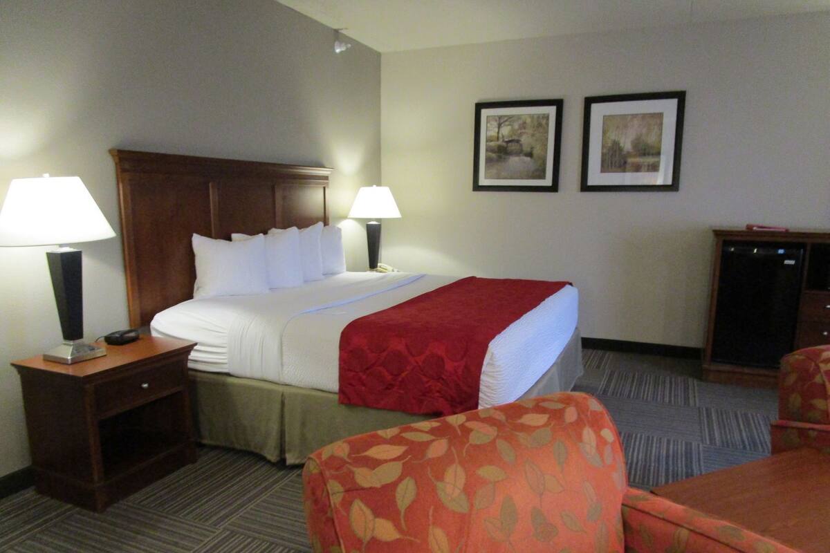 Une chambre d’hôtel avec un lit, deux lampes de chevet, une couverture rouge, une tête de lit en bois, deux tableaux encadrés au mur, un bureau avec un téléphone et deux fauteuils rembourrés.