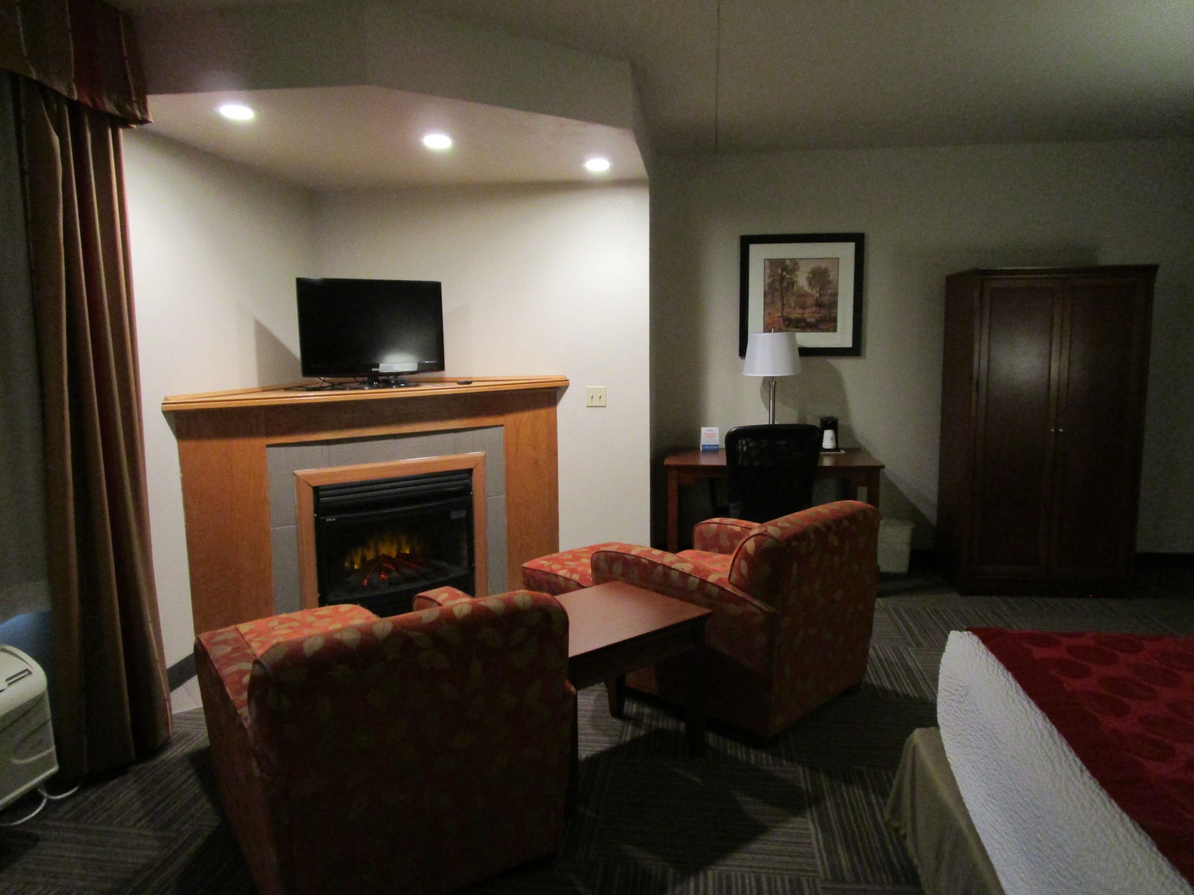 A hotel room with a fireplace, a television, a desk, two chairs, a bed, and a wardrobe.