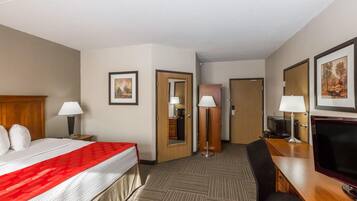 Habitación de hotel con una cama, dos mesitas de noche, un escritorio con una silla, un televisor, un espejo y dos puertas.