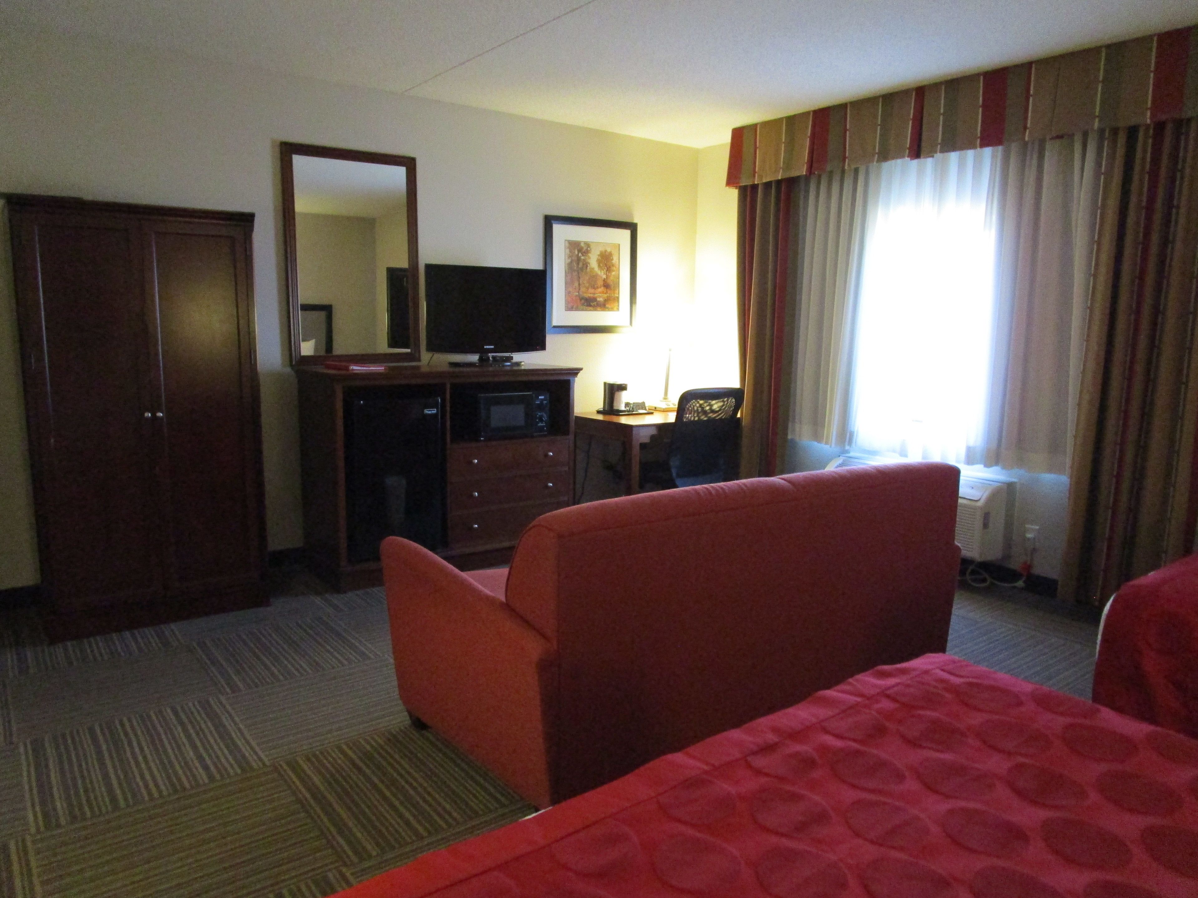 Quarto de hotel com cama, escrivaninha, televisão, guarda-roupa e janela com cortinas.