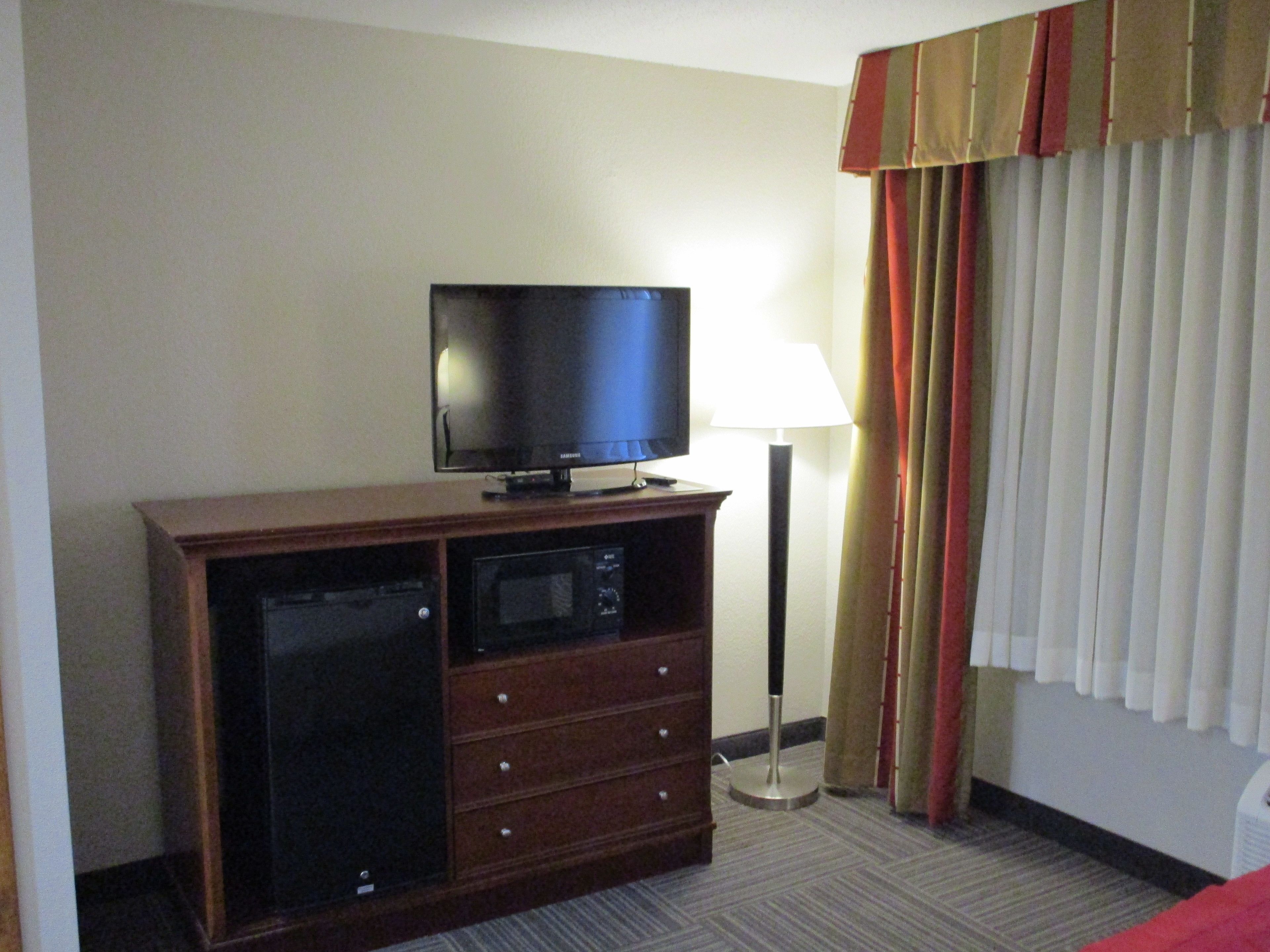 Habitación de hotel con televisor de pantalla plana sobre un mueble de madera, una lámpara y una ventana con cortinas.
