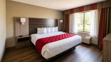 A hotel room with a large bed, two bedside tables, a nightstand, a window with curtains, and a wall-mounted heater.