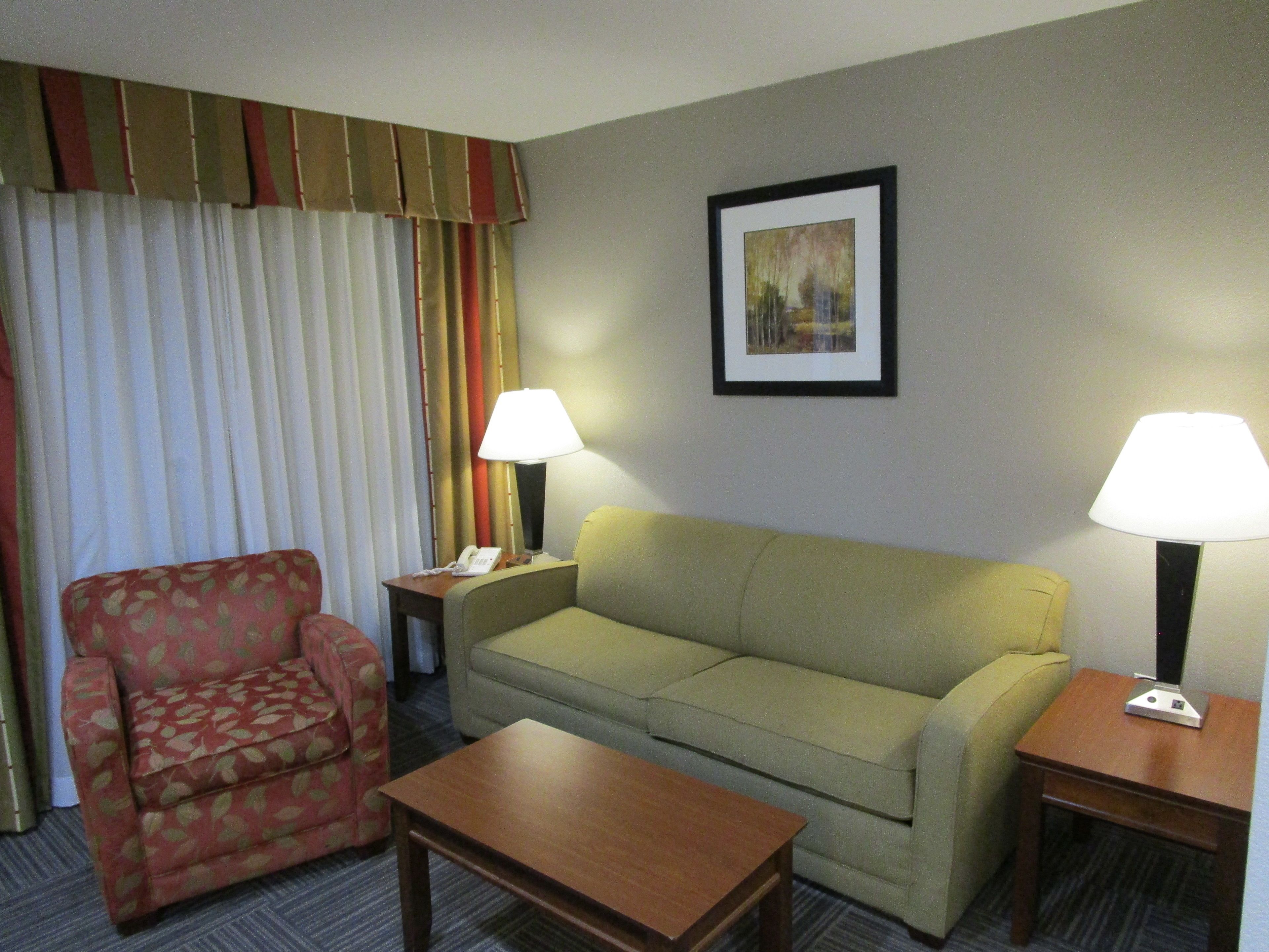 A hotel room with a sofa, a chair, a table, two lamps, a telephone, and a painting on the wall.