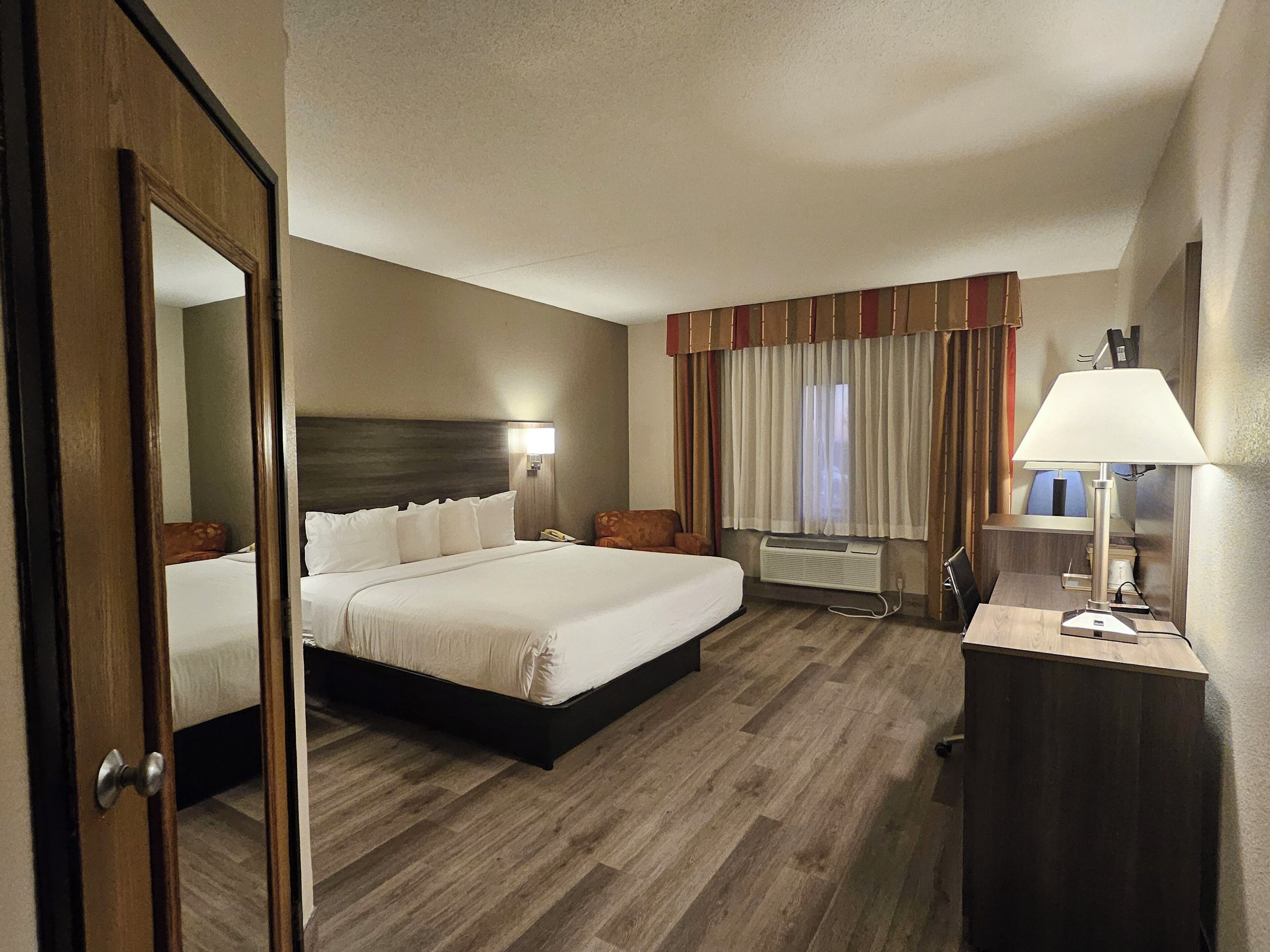 A hotel room with a large bed, a desk with a lamp, a bedside table, a mirror, a window with curtains, and an air conditioning unit.