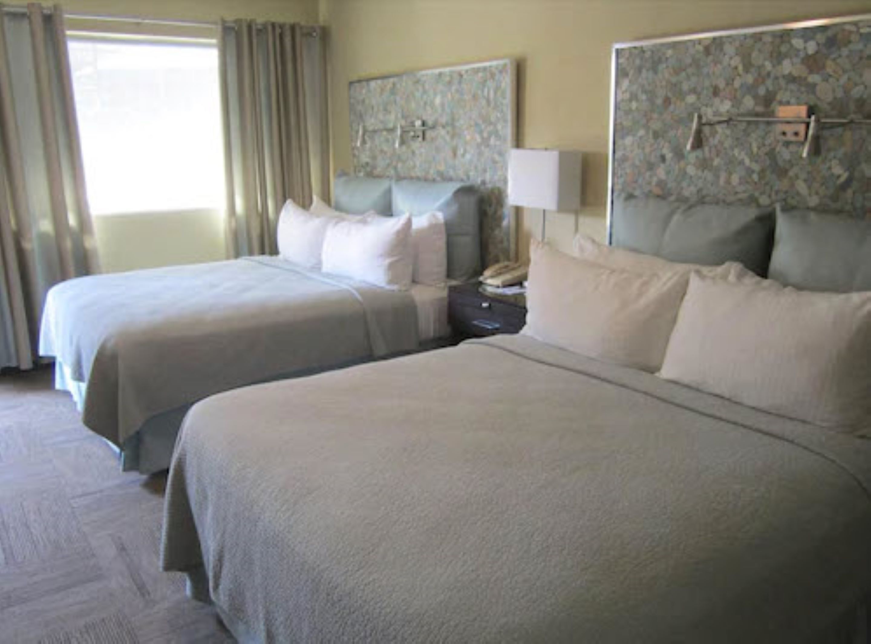 Una habitación de hotel con dos camas, una mesita de noche, una lámpara y un cabecero con motivos florales.